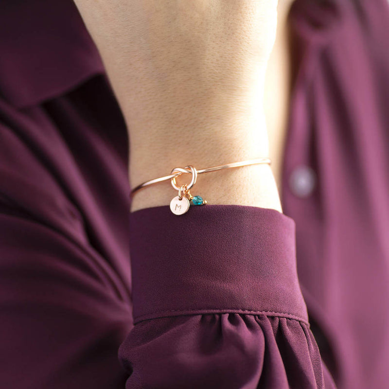 Model wears rose gold plated Friendship Knot Bangle with a small disc initial charm with the letter 'M' and a small blue zircon Swarovski birthstone charm for the month of December.