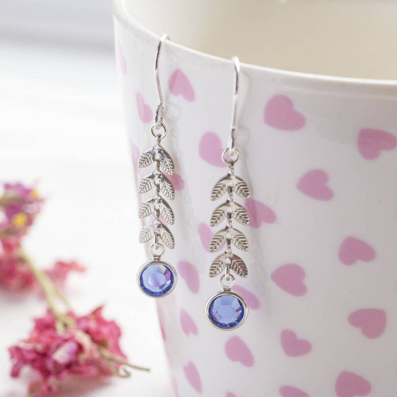 Image shows a pair of silver leaf vine earrings with September Sapphire Swarovski Birthstone detail. Earrings are hanging off a white cup with pink hearts.