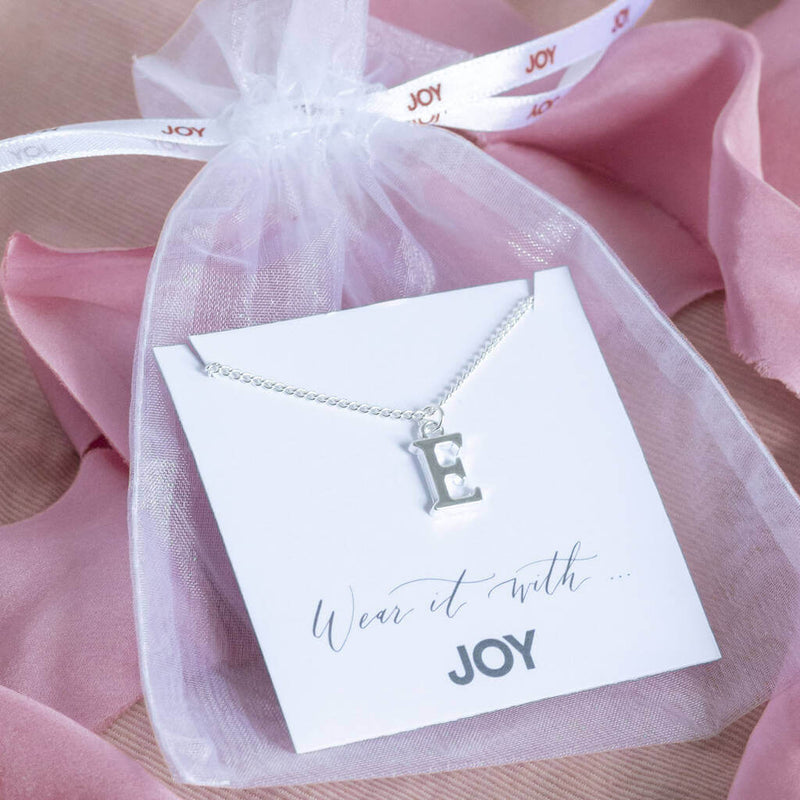 Image shows silver plated initial alphabet charm necklace with the letter 'E' on the 'Wear it with JOY' sentiment card in the white organza bag with 'JOY' ribbon. Organza bag and sentiment card free withe every purchase.