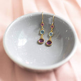 Gold plated Three Generations Birthstone Earrings. Birthstones in selection order from bottom to top: February Amethyst, March Aquamarine and October Rose. Earrings sit on a grey decorative bowl on a pink backdrop.