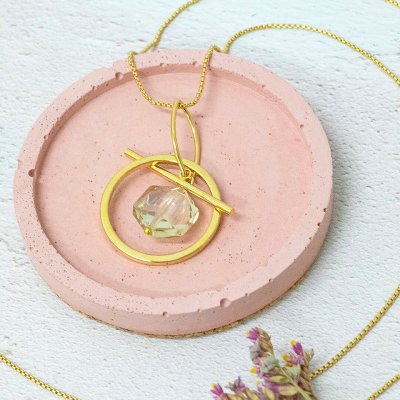 Image shows venus hexagonal necklace with a clear crystal hex within a golden circle pendant on a long length chain. Necklace sits on a pink background.