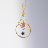 Image shows gold plated sunburst charm with a deep blue crystal attached, suspended within a golden arc.