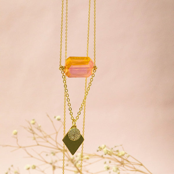 image shows apollo gold plated long length necklace featuring an orange crystal, hammered full moon and gold plated diamond shaped charm. Necklace suspended in front of a pink backdrop. 