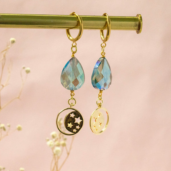 image shows celestial being huggie hoops with blue teardrop crystal and moon and stars charm.