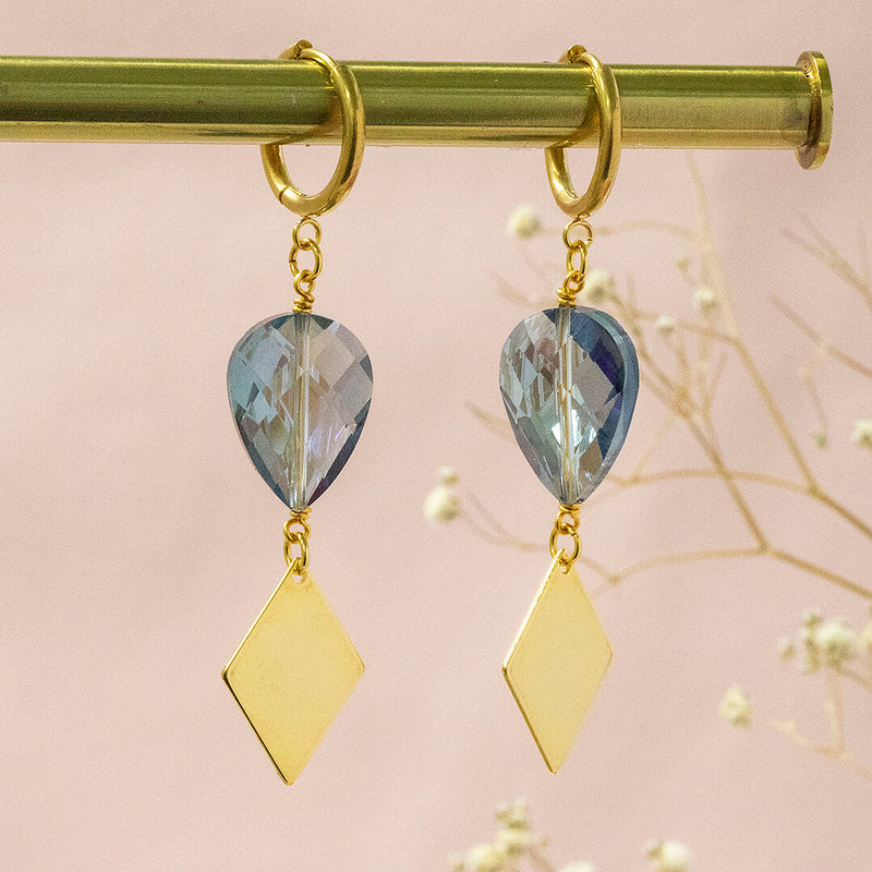 image shows a pair of diamonds in the sky earrings with blue tear shaped crystal completed with a gold plated diamond shaped charm. Suspended on a pink backdrop.