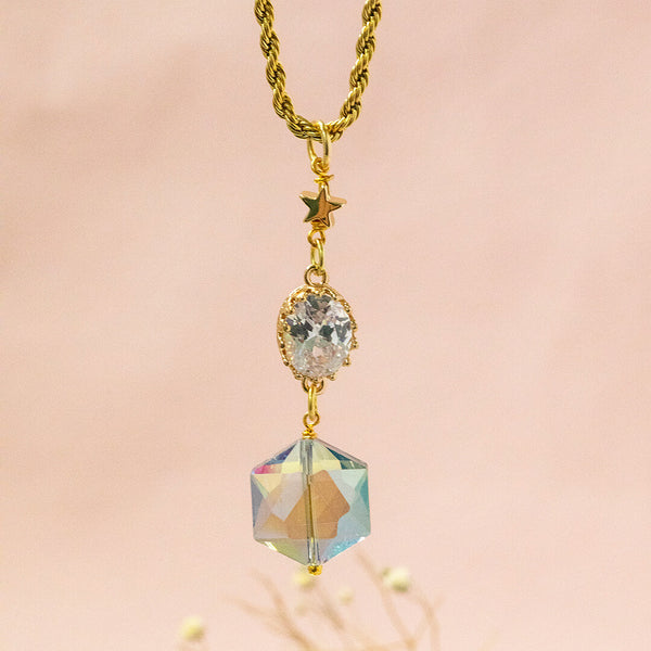 Image shows Elara gold plated necklace suspended in front of a pink backdrop. Necklace has hexagonal blue iridescent crystal with an oval clear crystal and a dainty gold plated star charm, complete with rope chain. 