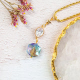 Image shows Elara gold plated necklace on a white backdrop. Necklace has hexagonal blue iridescent crystal with an oval clear crystal and a dainty gold plated star charm, complete with rope chain. 