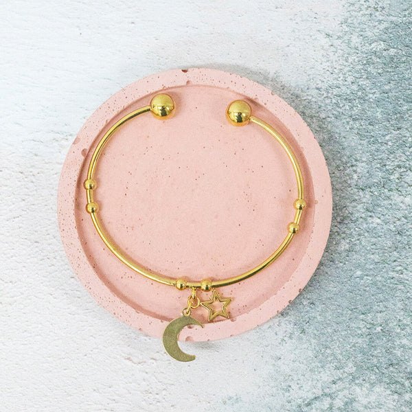 image shows gold plated heavenly bangle with crescent moon and star charms sat on a pink backdrop.