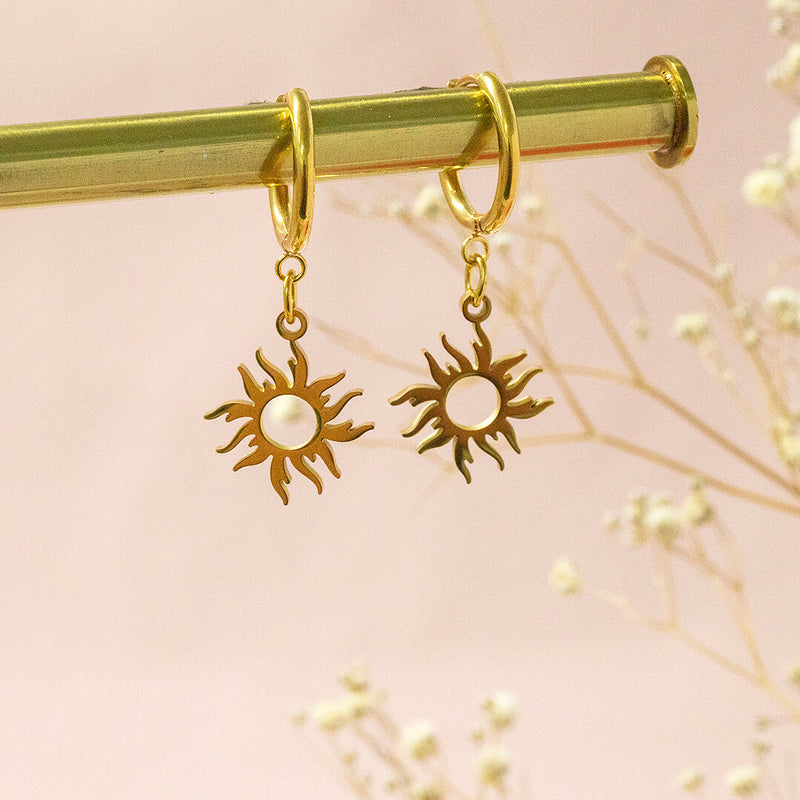 Helios gold plated earrings with sunburst hanging in front of a pink backdrop.