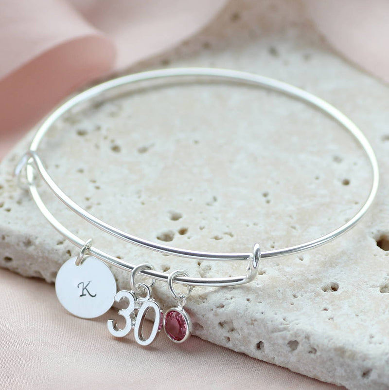 Image shows milestone birthday bangle with initial charm with the letter 'k', '30' number charm and October Rose Swarovski crystal birthstone charm.