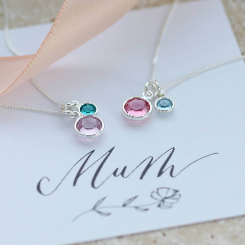 image shows two sterling silver mother and child birthstone charm necklaces from the left, necklace has June light amethyst and May emerald. Second necklace has October Rose and March aquamarine Swarovski crystal charms, flat lay on 'Mum' sentiment card.