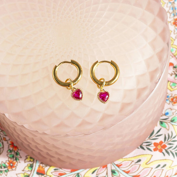 Image shows gold plated hoop earrings with July Ruby birthstone heart charms placed on a pink trinket dish