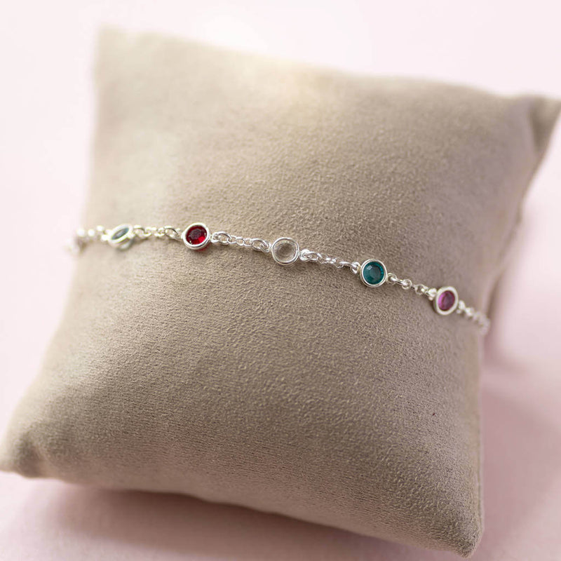 Image shows Sterling silver mini family birthstone bracelet with six birthstones (up to eight can be selected) from right to left birthstones are October Rose, May Emerald, April Crystal, July Ruby, March Aquamarine and February Amethyst wrapped around a jewellery pillow.