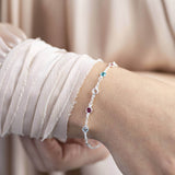 Model wears sterling silver mini family birthstone bracelet with five visible birthstones (up to eight can be selected) from top to bottom birthstones are October Rose, May Emerald, April Crystal, July Ruby, March Aquamarine,