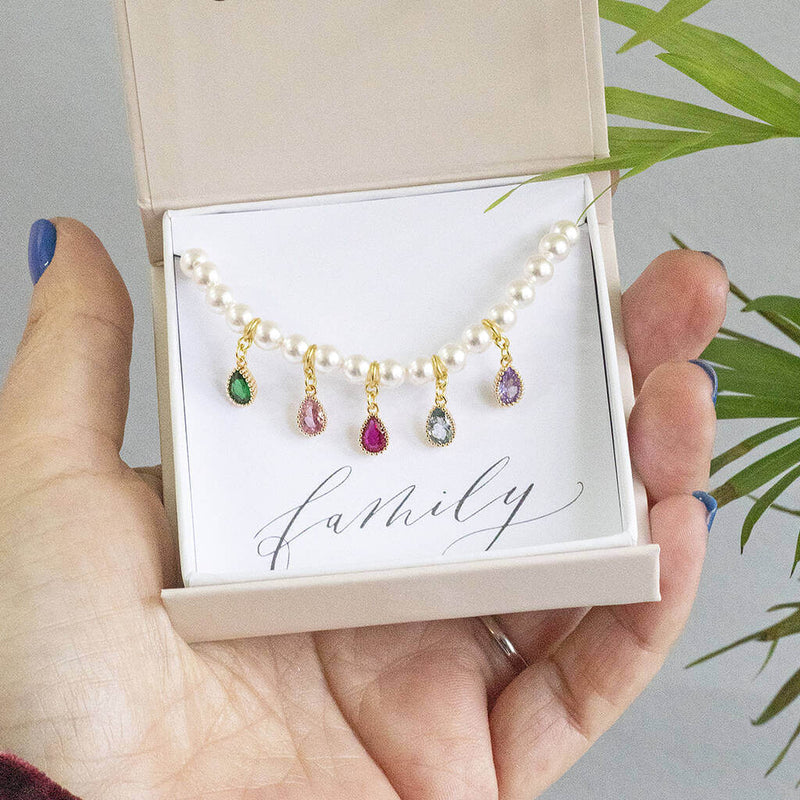 image shows pearl bracelet with family birthstone teardrops. Five birthstones appear here to represent different family members displayed on a 'family' sentiment card in a JOY by Corrine Smith gift box.