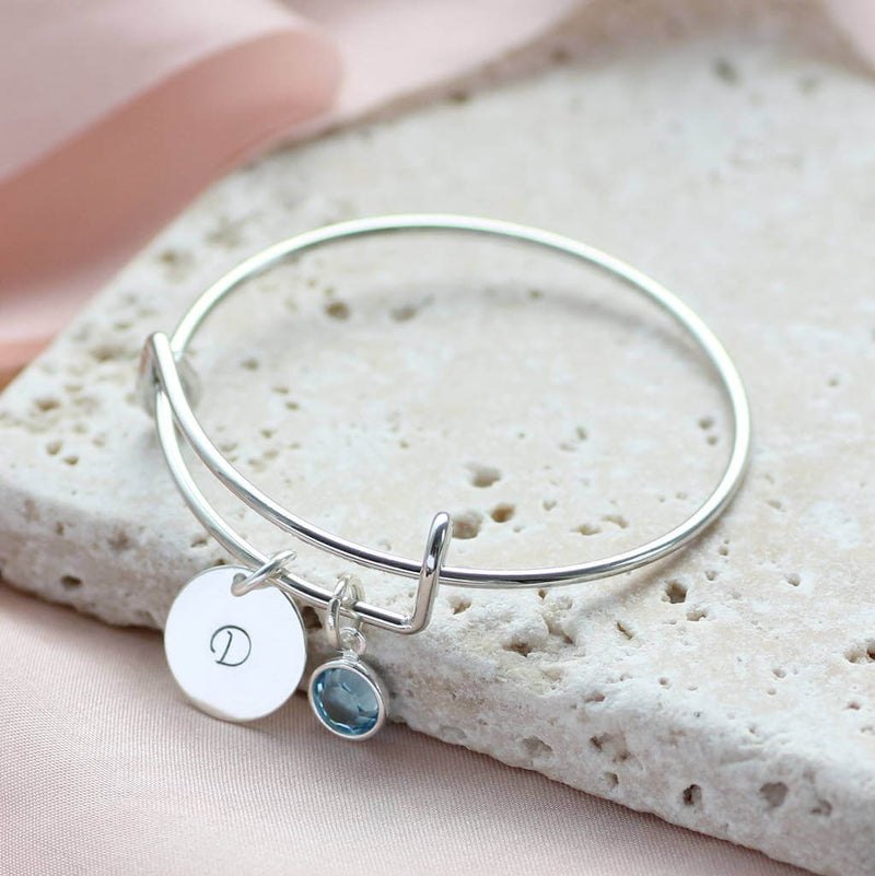 Image shows personalised birthstone christening bangle with Swarovski birthstone and hand stamped initial disc with the letter 'D'