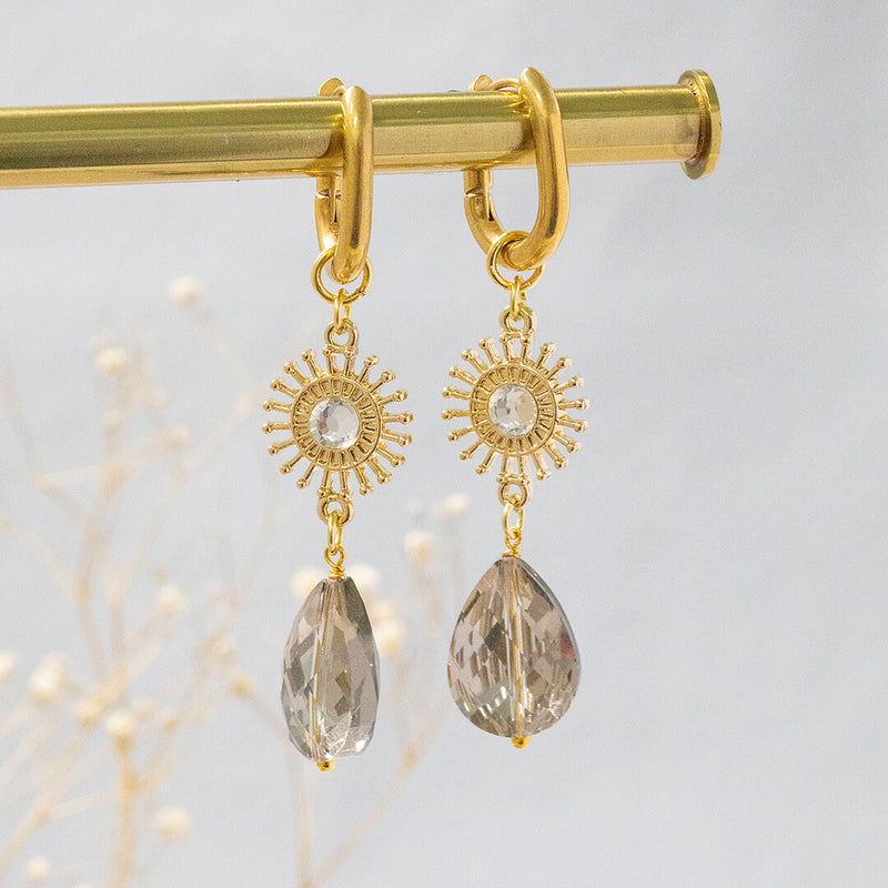 image shows radiance sunburst earrings with grey crystal and sunburst charm on a grey background.