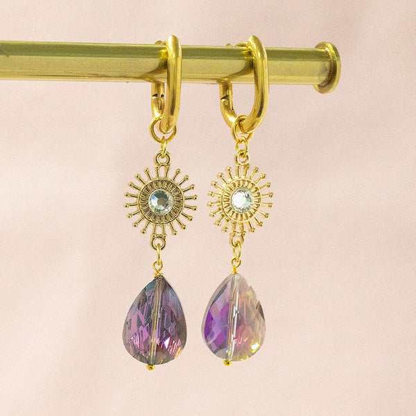Image shows a pair of radiance sunburst purple crystal earrings suspended in front of a pink backdrop, 