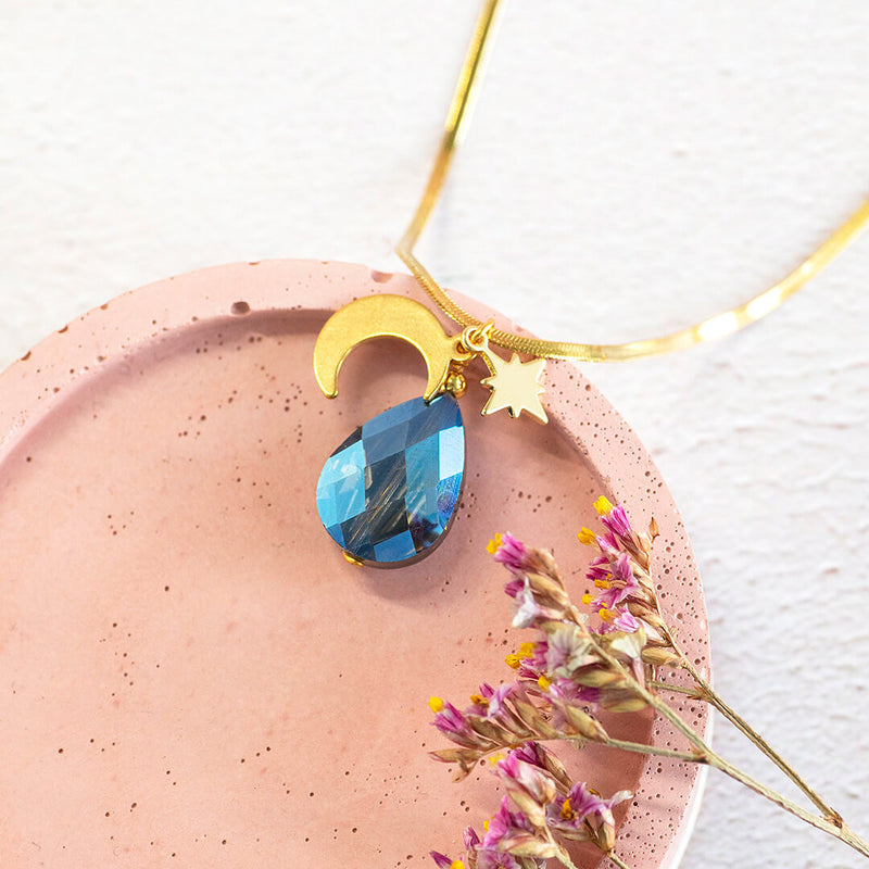 Image shows selene moon and star gold plated pendant with blue teardrop crystal. On a pink and grey background.