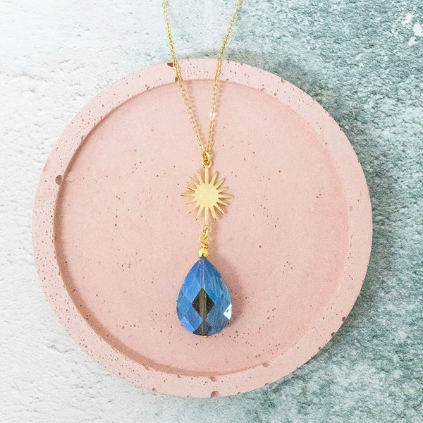 Image shows a solaris blue crystal necklace with sunburst charm on a pink backdrop