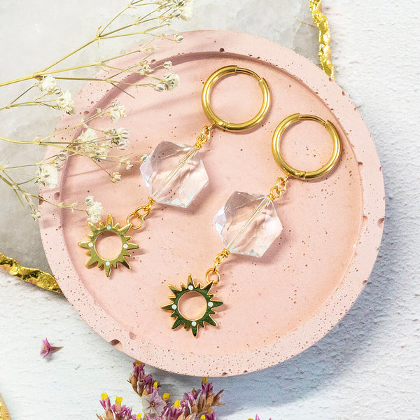 Image shows soleil gold plated earrings sat on a pink backdrop and tiny pink and white flowers. Earrings feature clear hexagonal crystal and tiny sunburst charm embedded with opals.  