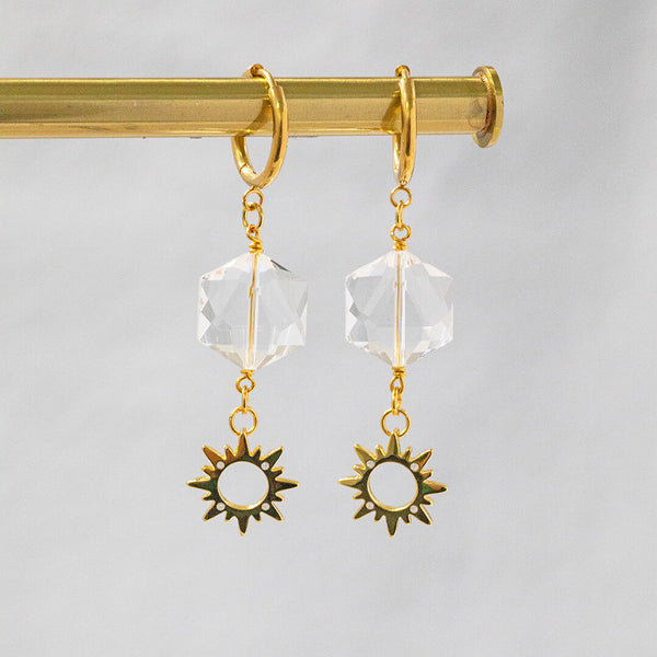 Image shows soleil gold plated earrings suspended in front of a white backdrop. Earrings feature clear hexagonal crystal and tiny sunburst charm embedded with opals.  