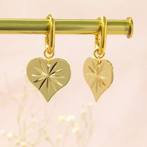 Starstruck gold plated heart earrings suspended in front of a pink backdrop.